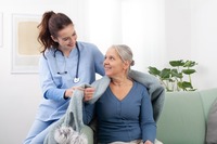 Das Bild zeigt eine Pflegerin, die einer älteren Frau zu Hause eine Decke um die Schultern.